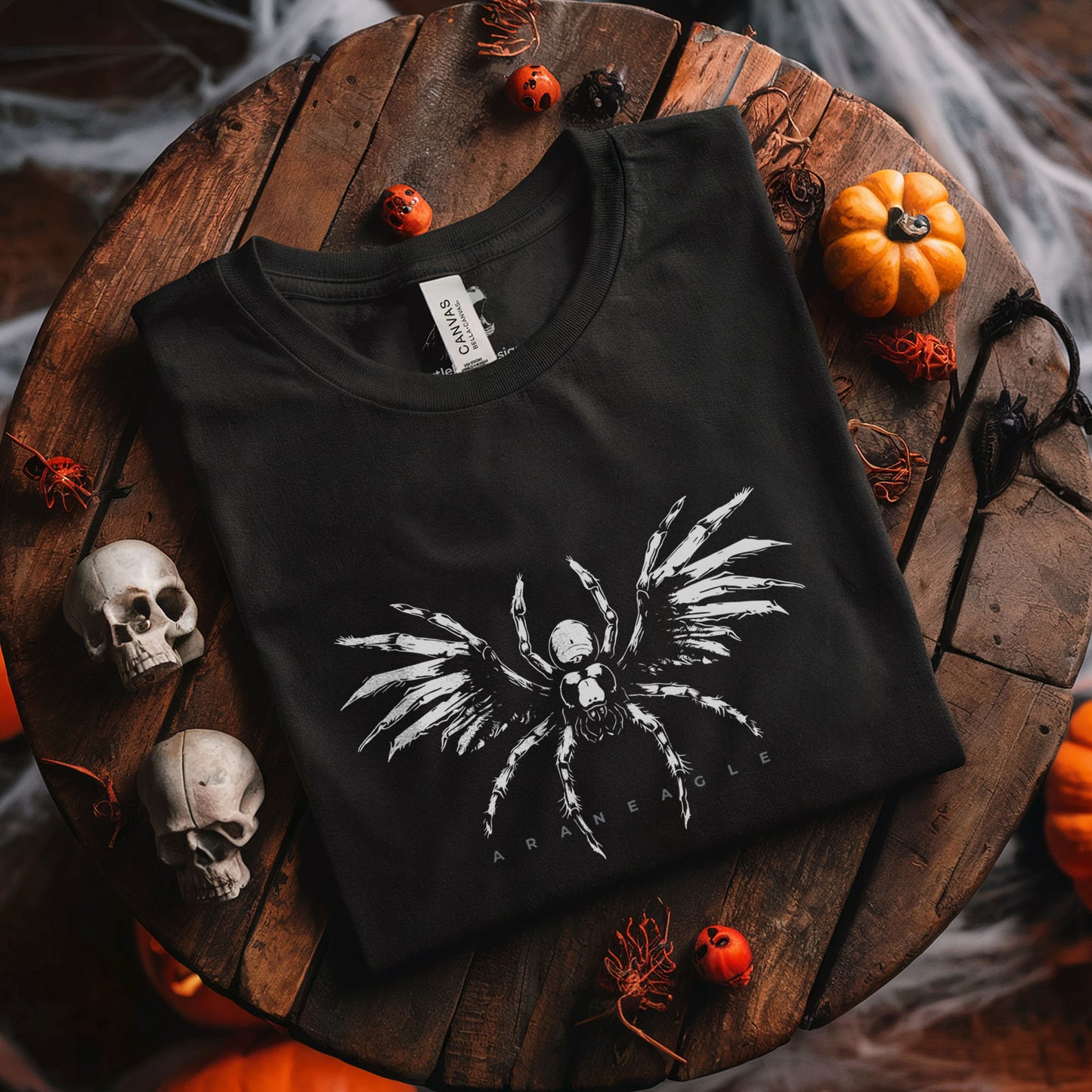 A black short-sleeve graphic T-shirt laid flat on a rustic wooden surface, surrounded by Halloween props including skulls, small pumpkins, spiderwebs, and orange berries. The shirt has a large white skeletal spider design with the word 'ARANEAGLE' printed below it.