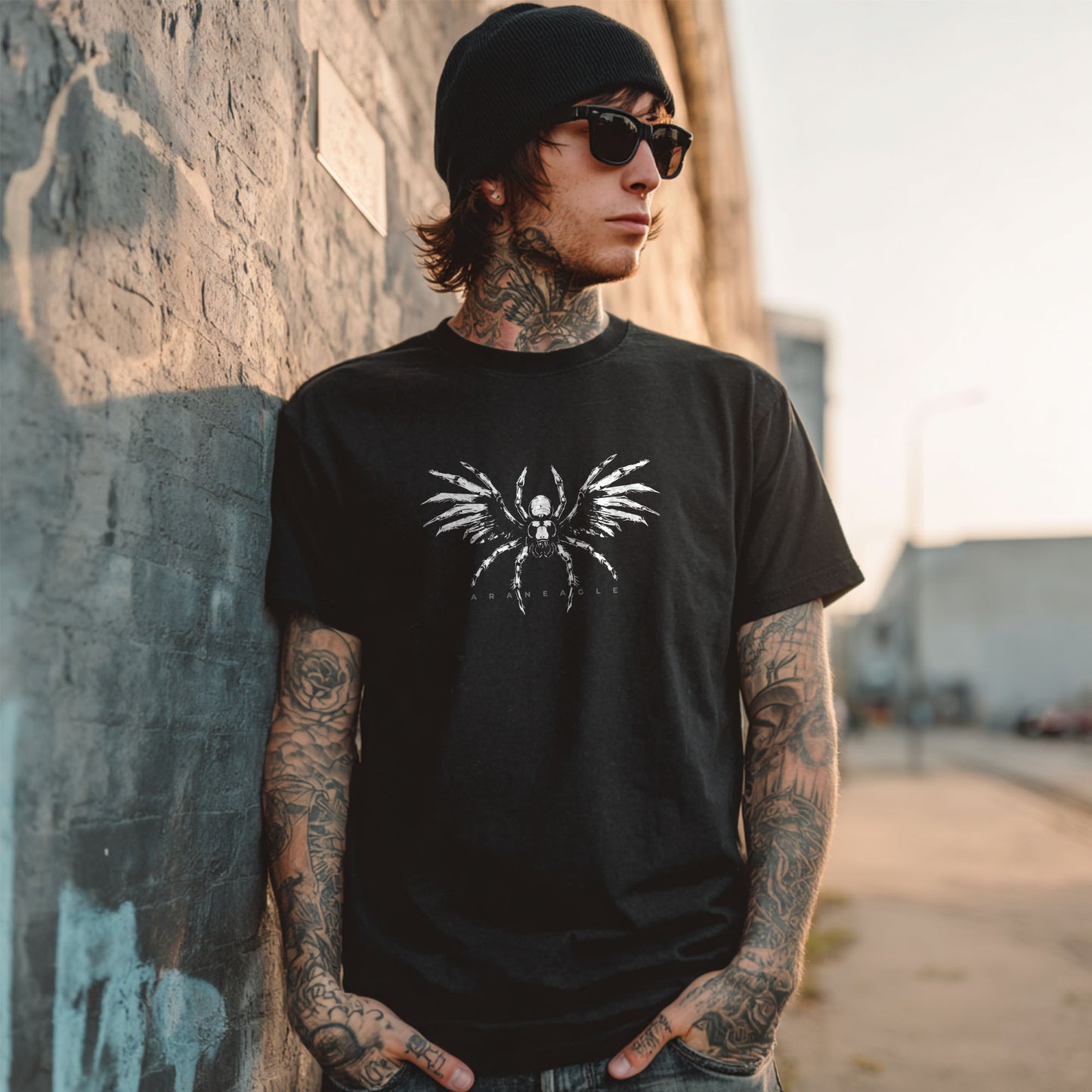 A tattooed man wearing a black beanie, sunglasses, and a black graphic t-shirt with a white spider design, leaning against a concrete wall in an outdoor urban setting.